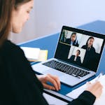 A woman engaging in a video conference using a laptop at home, taking notes.